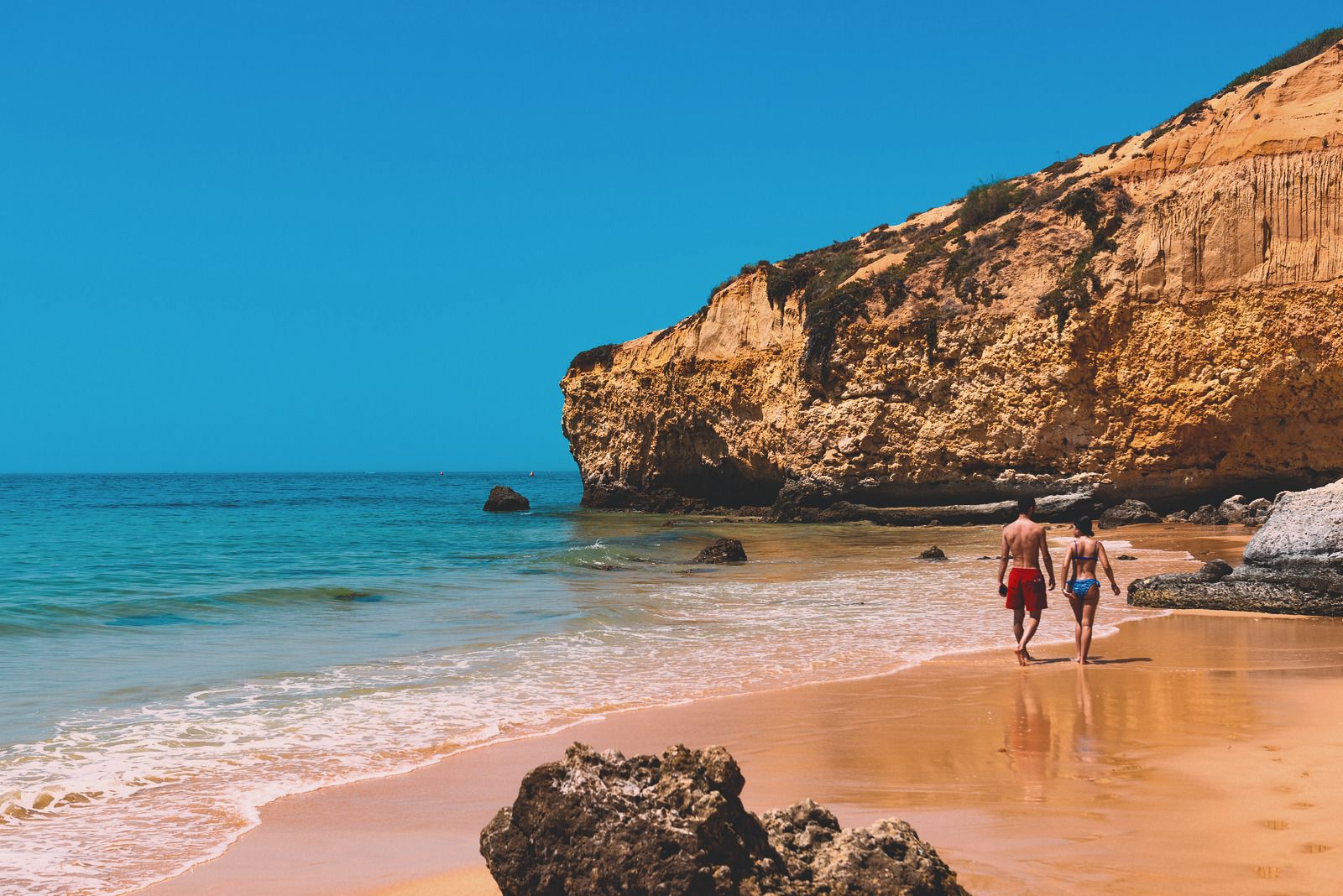 Zwei Menschen gehen an der Wasserlinie eines ruhigen Algarve-Strands unter goldenen Klippen entlang