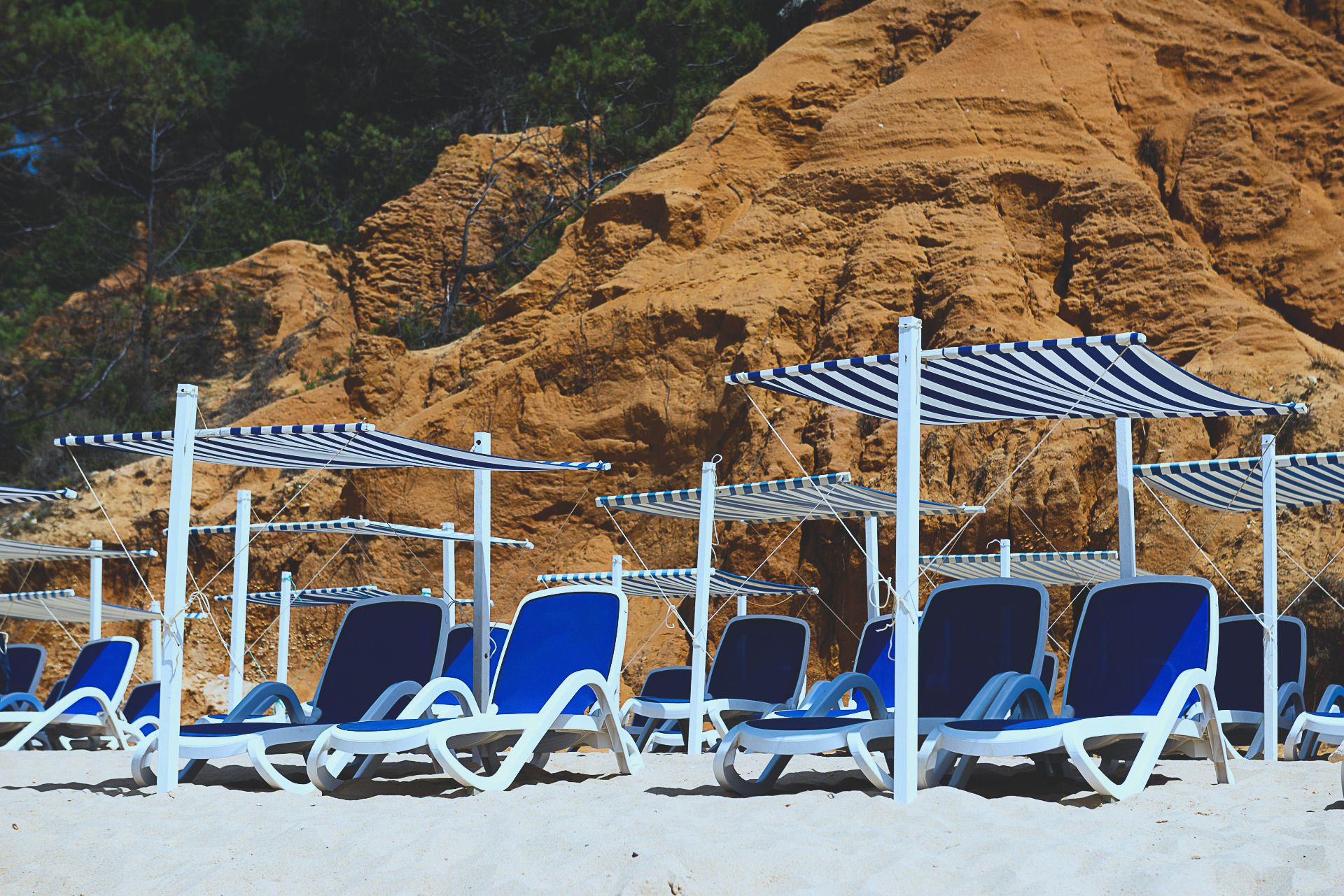 Gestreifte Sonnenschirme und blaue Liegen im Sand unter einer ockerfarbenen Klippe